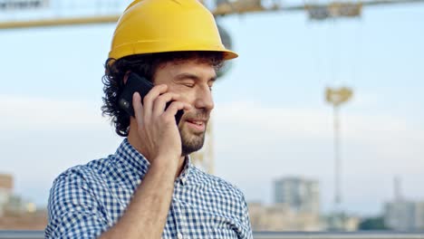 Close-up-of-the-young-Caucasian-handsome-man-in-the-yellow-hardhat-talking-on-the-phone-and-smiling-at-the-building-site.-Outdoor.